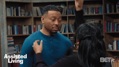 A Woman Is Touching A Man 'S Face In Front Of A Bookshelf With Assisted Living Written On It GIF