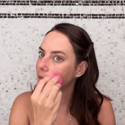 A Woman Is Using A Pink Sponge To Apply Makeup To Her Face GIF