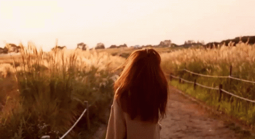 A Woman Is Walking Down A Dirt Road In A Field . GIF