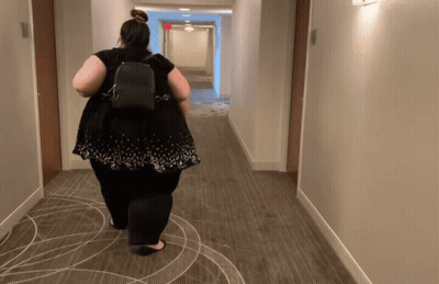 A Woman Is Walking Down A Hallway With A Backpack GIF