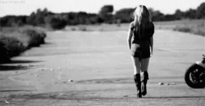 A Woman Is Walking Down A Road In Front Of A Motorcycle . GIF