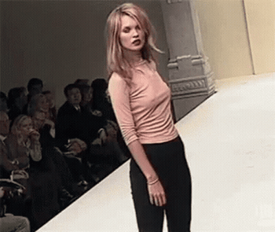 A Woman Is Walking Down A Runway At A Fashion Show Wearing A Pink Shirt And Black Pants . GIF