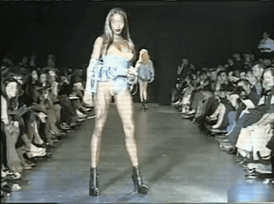 A Woman Is Walking Down A Runway At A Fashion Show While A Crowd Watches . GIF