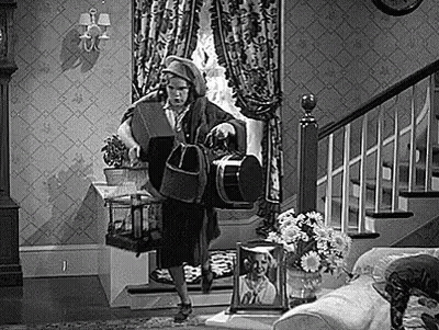 A Woman Is Walking Down A Set Of Stairs In A Living Room Holding A Dog . GIF