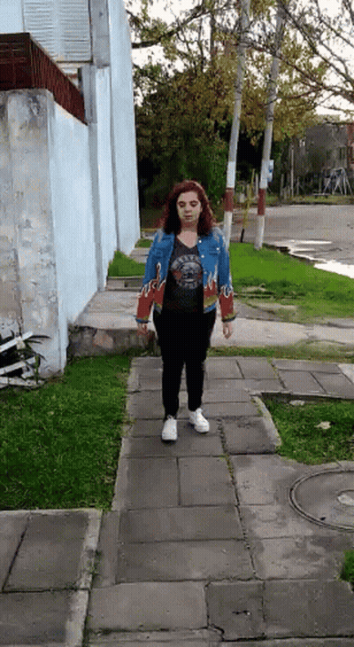 A Woman Is Walking Down A Sidewalk Wearing A Blue Jacket With Flames On The Sleeves GIF