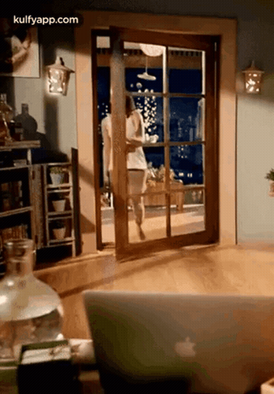 A Woman Is Walking Through A Sliding Glass Door In A Living Room Next To An Apple Laptop . GIF