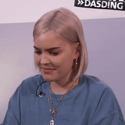 A Woman Is Wearing A Blue Shirt And A Necklace With A Letter A On It GIF