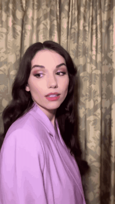 A Woman Is Wearing A Purple Jacket And Standing In Front Of A Curtain GIF