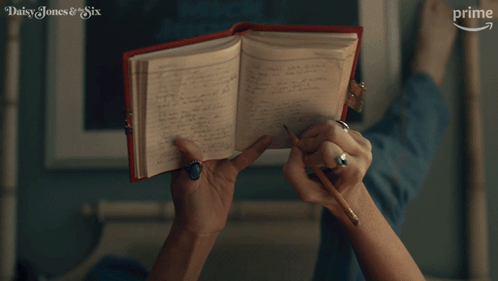 A Woman Is Writing In A Notebook With The Words Daisy Jones & Six On The Bottom Right GIF