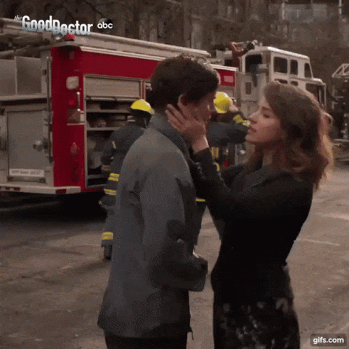 A Woman Kisses A Man On The Cheek In Front Of A Fire Truck That Says The Good Doctor On It GIF