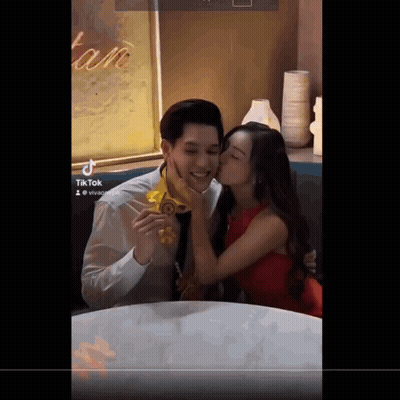 A Woman Kisses A Man On The Cheek In Front Of A Sign That Says " Tan " GIF