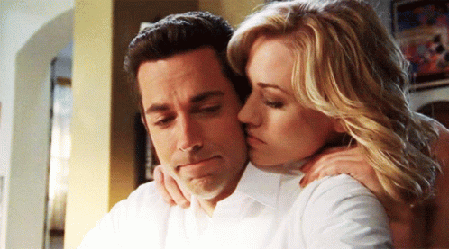 A Woman Kisses A Man On The Cheek While He Looks Down GIF