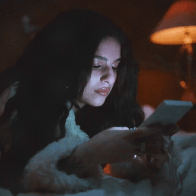A Woman Laying On A Bed Looking At A Cell Phone GIF