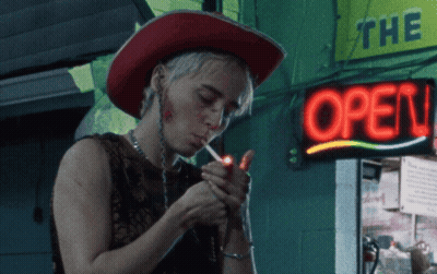 A Woman Lighting A Cigarette In Front Of A Neon Sign That Says Open GIF