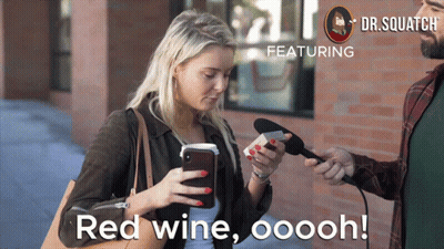 A Woman Looks At Her Phone While A Man Holds A Microphone And Says Red Wine Ooooh GIF