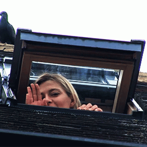 A Woman Looks Out Of A Window With Her Hand In Front Of Her Face GIF