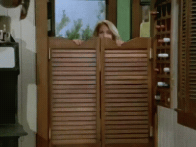 A Woman Peeking Out From Behind A Wooden Saloon Door GIF
