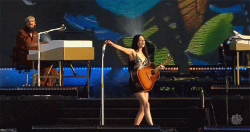 A Woman Playing A Guitar On Stage With A Man Playing A Keyboard In The Background GIF