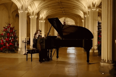 A Woman Playing A Piano In A Hallway With Christmas Trees GIF