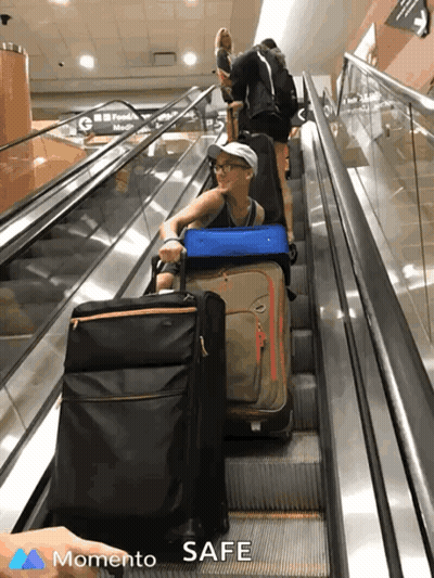 A Woman Pushes Her Luggage Up An Escalator With The Words " Momento Safe " On The Bottom Right GIF