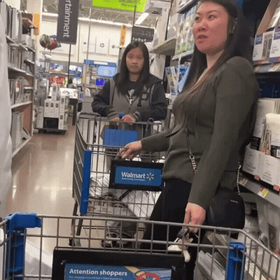 A Woman Pushing A Shopping Cart In A Store With A Sign That Says Attention Shoppers GIF