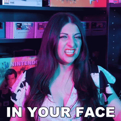 A Woman Says In Your Face While Standing In Front Of A Shelf Full Of Video Games GIF