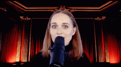A Woman Singing Into A Microphone In Front Of A Red Curtain GIF