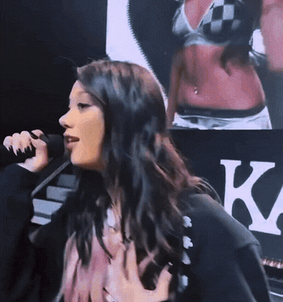 A Woman Singing Into A Microphone In Front Of A Sign That Says K. GIF