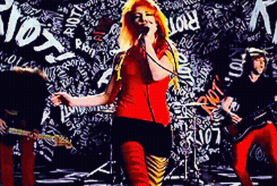 A Woman Singing Into A Microphone In Front Of A Wall With The Word Riot Written On It GIF