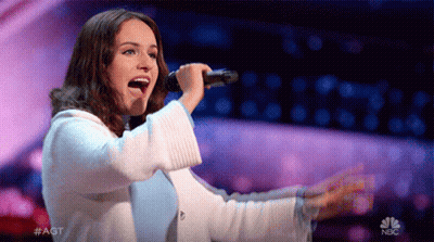 a woman singing into a microphone on a stage with a nbc logo in the background