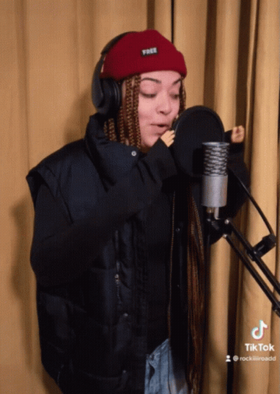 A Woman Singing Into A Microphone Wearing Headphones And A Red Beanie That Says Free GIF