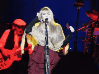 A Woman Singing Into A Microphone While A Man Plays A Guitar In The Background GIF