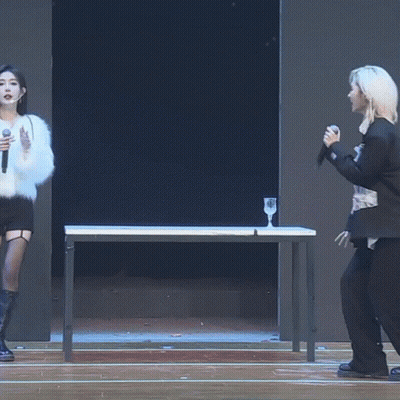 A Woman Singing Into A Microphone While Standing Next To A Table GIF