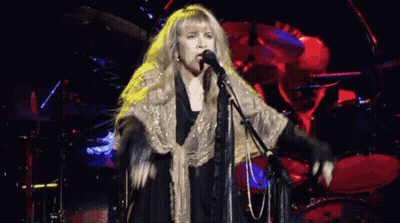 A Woman Singing Into A Microphone With A Drum Set In The Background GIF