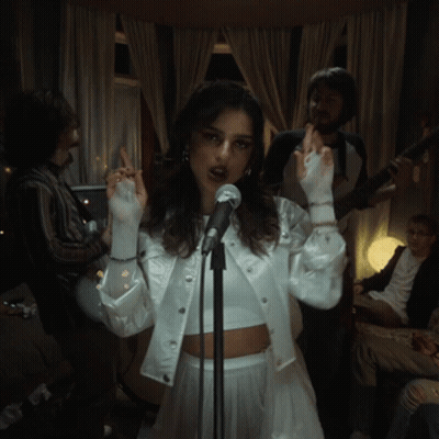 A Woman Singing Into A Microphone With A Man Playing A Guitar Behind Her GIF