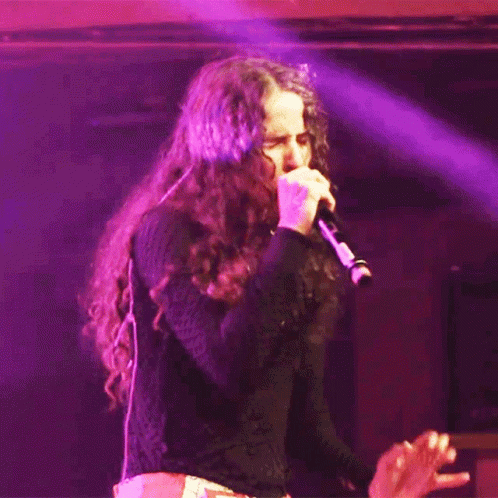 A Woman Singing Into A Microphone With Purple Lights Behind Her GIF