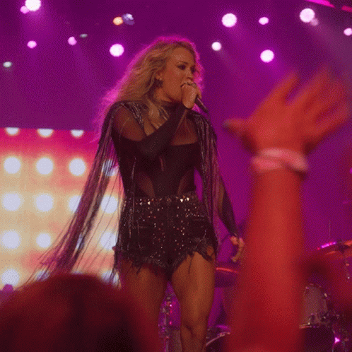 A Woman Singing Into A Microphone With Purple Lights In The Background GIF
