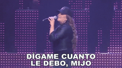 A Woman Singing Into A Microphone With The Words " Digame Cuanto Le Debo Mijo " Behind Her GIF
