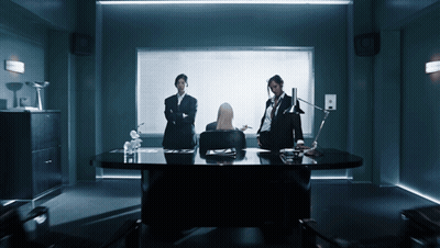A Woman Sits At A Desk In A Dark Room With Two Men Standing Around Her GIF