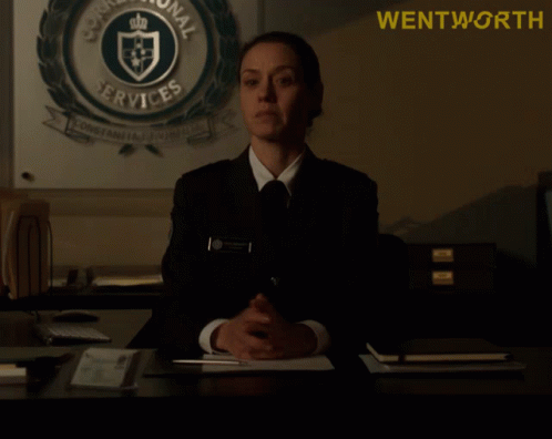 A Woman Sits At A Desk In Front Of A Sign That Says " Wentworth " GIF