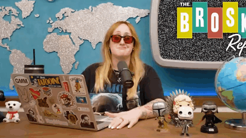 A Woman Sits At A Desk With A Laptop And A Microphone In Front Of A Sign That Says The Broski Reg GIF
