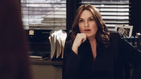 A Woman Sits At A Desk With Her Hand On Her Chin And A Picture Of A Police Officer Behind Her GIF