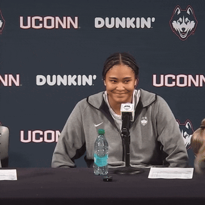 A Woman Sits At A Table In Front Of A Wall That Says Dunkin Uconn GIF