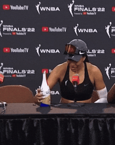 A Woman Sits At A Table In Front Of A Wall That Says Finals 22 On It GIF