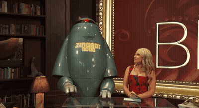 A Woman Sits At A Table Next To A Robot That Says Zioshot 9000 On It GIF