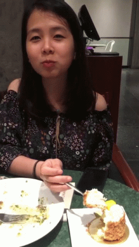 A Woman Sits At A Table With A Plate Of Food And A Piece Of Cake GIF
