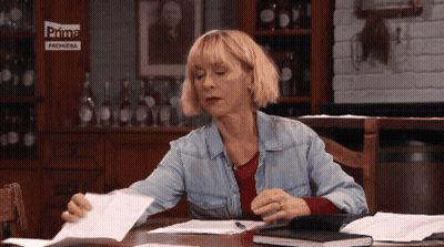 A Woman Sits At A Table With Papers In Front Of A Sign That Says Prima Premiera GIF