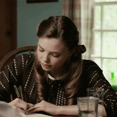 A Woman Sits At A Table Writing With A Pen GIF