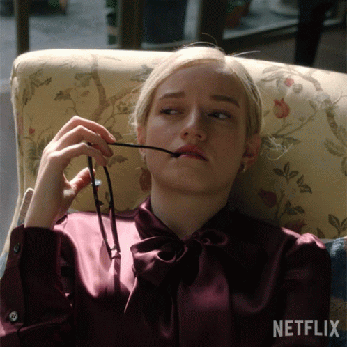 A Woman Sits In A Chair Holding A Pair Of Glasses In Her Mouth GIF
