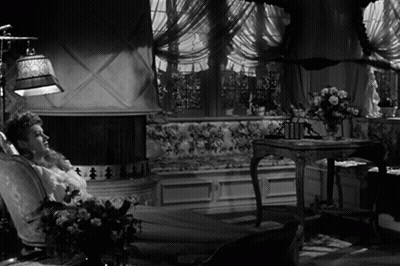 A Woman Sits In A Chair In Front Of A Fireplace In A Black And White Photo GIF